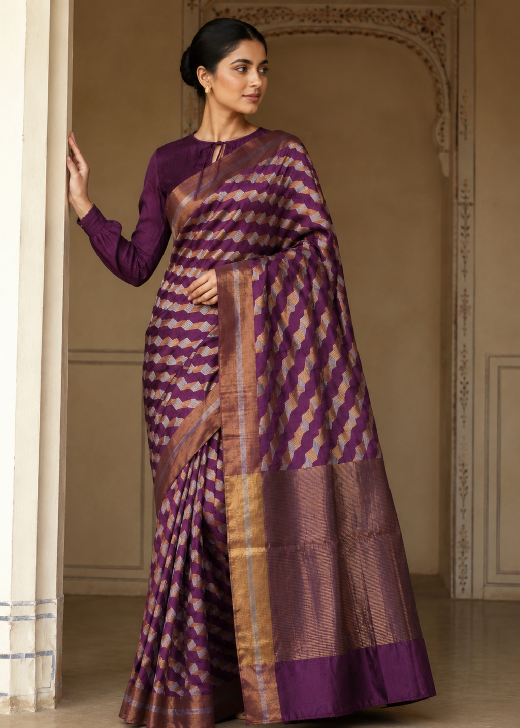 A woman models a purple and gold patterned silk saree with a matching purple long-sleeve blouse in an elegant indoor setting with beige walls and arches.