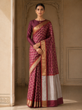 A woman models a traditional Indian saree in a grand, historic interior with beige stone arches. The saree is a deep maroon/burgundy with a gold geometric lattice pattern, a solid maroon blouse, and a wide silver and gold border.