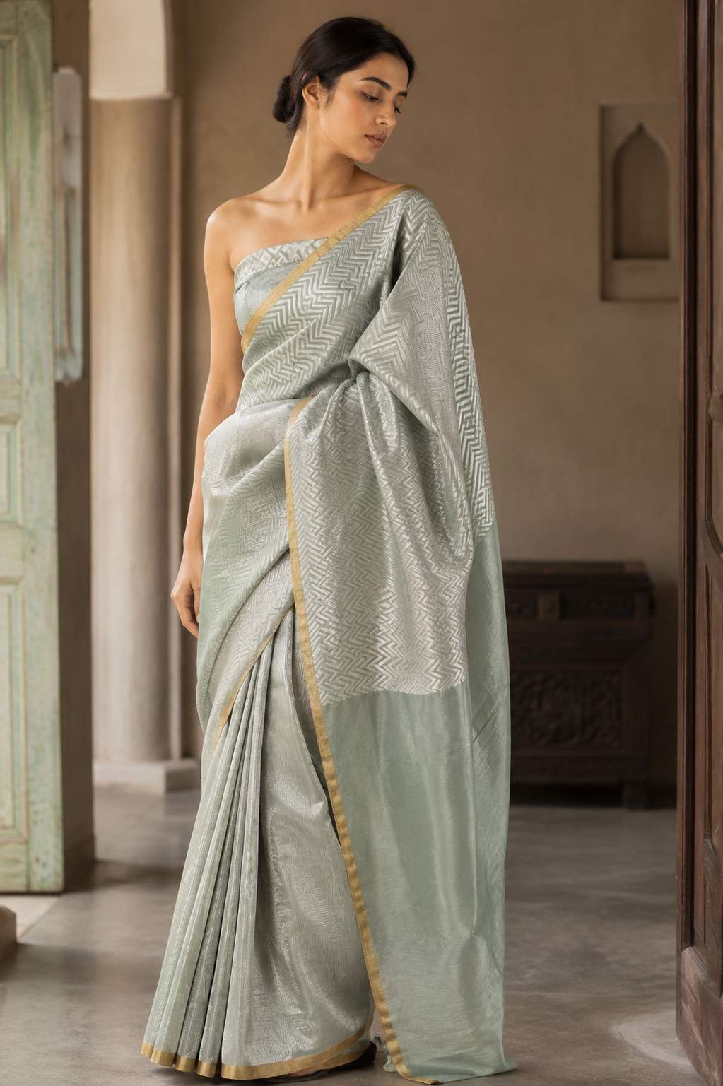 A woman wearing a sage green silk saree with a silver zari geometric pattern and a gold border, standing in a traditional indoor setting.