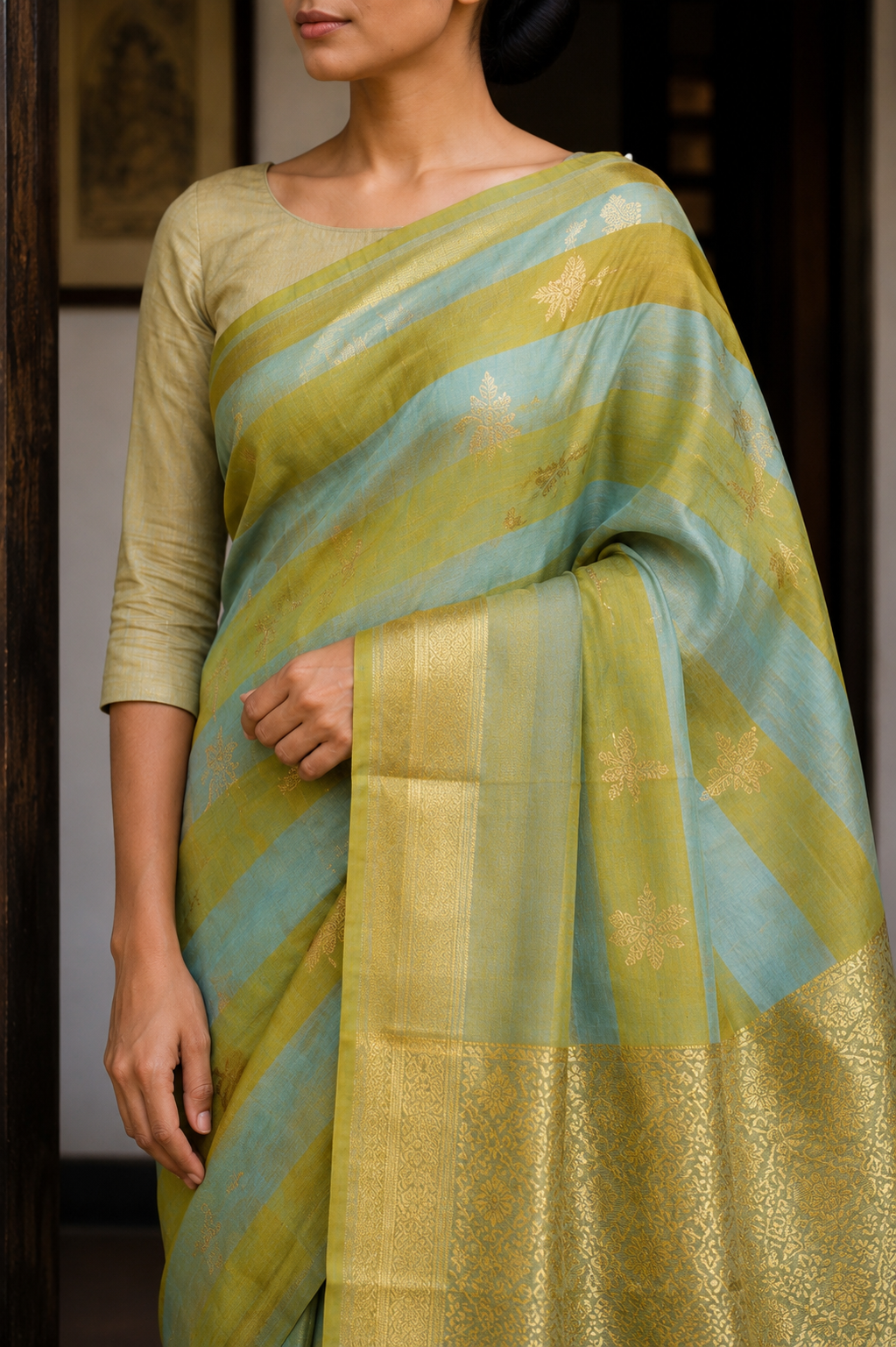 A woman wearing a light olive green and light blue striped silk saree with gold zari floral motifs and a wide gold border, paired with a matching olive green blouse.