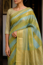 A woman wearing a light olive green and light blue striped silk saree with gold zari floral motifs and a wide gold border, paired with a matching olive green blouse.