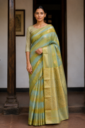 A woman wearing a light green and gold striped silk saree with a gold zari border, standing in front of a dark doorway.